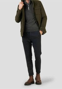 Giacca verde oliva sopra un maglione grigio, abbinata a pantaloni scuri dai tessuti strutturati e stivaletti marroni alla caviglia. La cerniera argentata sulla giacca aggiunge un tocco di dettaglio.