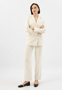 Abito crema a doppio petto con vestibilità su misura, pantaloni dritti e ballerine nere con un sottile accentuato metallico. Tessuto morbido e design strutturato.