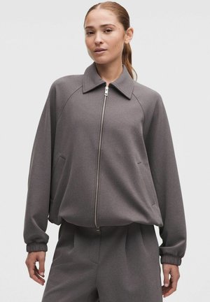 Mujer con chaqueta gris con cremallera, cuello y mangas largas, combinada con pantalones grises a juego, sobre un fondo liso.