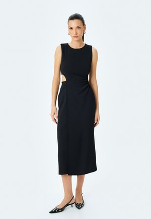 Schwarzes Midikleid mit ärmellosem Design, asymmetrischem Ausschnitt an der Taille, glatter Textur und minimalistischer Silhouette. Kombiniert mit verzierten Absätzen.