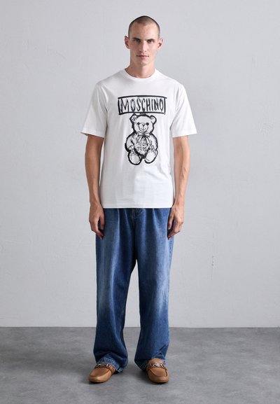 Vit bomullströja med en svarttryckt nallebjörn grafisk design och "MOSCHINO" text ovanför. Parat med blå denimbyxor och bruna skor.