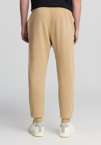 Pantaloni felpa beige con fascia elastica in vita e polsini alla caviglia, dotati di tasche laterali e una texture morbida. Indossati con scarpe da ginnastica bianche.