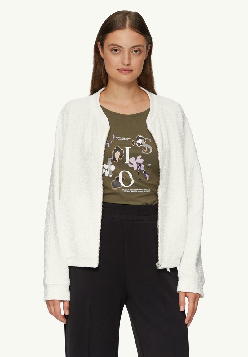 Weiße, strukturierte Bomberjacke mit Reißverschluss, die über einem olivgrünen Grafik-T-Shirt getragen wird. Das Modell posiert mit entspannten Ärmeln und schwarzen Hosen.