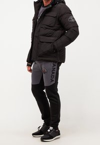 Schwarze gepolsterte Jacke mit mehreren Taschen und Kapuze, kombiniert mit grauen und schwarzen Sweatpants mit Kordelzug und Logo. Schwarze Sneaker runden das Outfit ab.