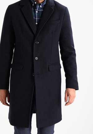 Homme portant un manteau en laine bleu marine foncé boutonné, avec une chemise à carreaux bleue et verte en dessous et un jean foncé.