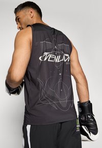 Haut de sport sans manches noir avec un design graphique blanc, présentant des lignes géométriques et le logo de la marque au dos. Tissu texturé et léger.