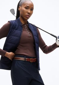 Γυναίκα με καφέ πουκάμισο μακρυμάνικο και μπλε γιλέκο, κρατώντας μπαστούνι γκολφ στον ώμο, φοράει μπλε παντελόνι και δερμάτινη ζώνη.