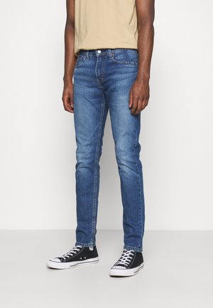 Jeans de denim ajustados en lavado azul medio. Presentan un diseño clásico de cinco bolsillos, con herrajes plateados y un ligero desgaste en las rodillas.