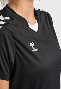 Svart atletisk t-shirt med v-ringning och korta ärmar. Har vita ränder på axlarna och en vit logotyp på bröstet. Slät tygstruktur.