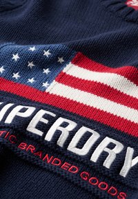 Tessuto a maglia navy con un design prominente della bandiera americana, caratterizzato da strisce rosse e bianche e stelle bianche, insieme a un marchio ricamato.