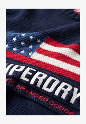 Tessuto a maglia navy con un design prominente della bandiera americana, caratterizzato da strisce rosse e bianche e stelle bianche, insieme a un marchio ricamato.