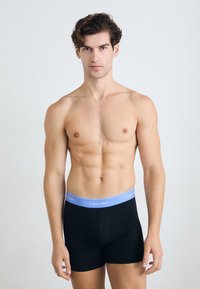 Pantaloni boxer neri con una cintura logo Calvin Klein azzurro chiaro. Il modello ha una corporatura atletica e si trova di fronte a uno sfondo neutro.