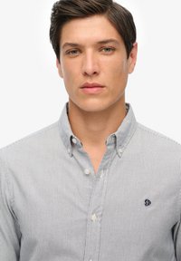 Jongeman met kort, donker haar, gekleed in een lichtgrijs en wit gestreept overhemd met knopen en een klein geborduurd logo op de borst.