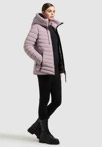 Kvinde iført en lys lilla polstret hættejakke over sorte leggings og sorte snørede kampstøvler, stående mod en ensfarvet baggrund.