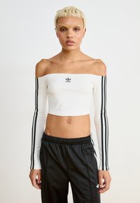 Bijela kratka majica s dugim rukavima crnih pruga, s otkrićem ramena i malim crnim adidas logom na prsima. Glatka tekstura, prilagodljiv dizajn.