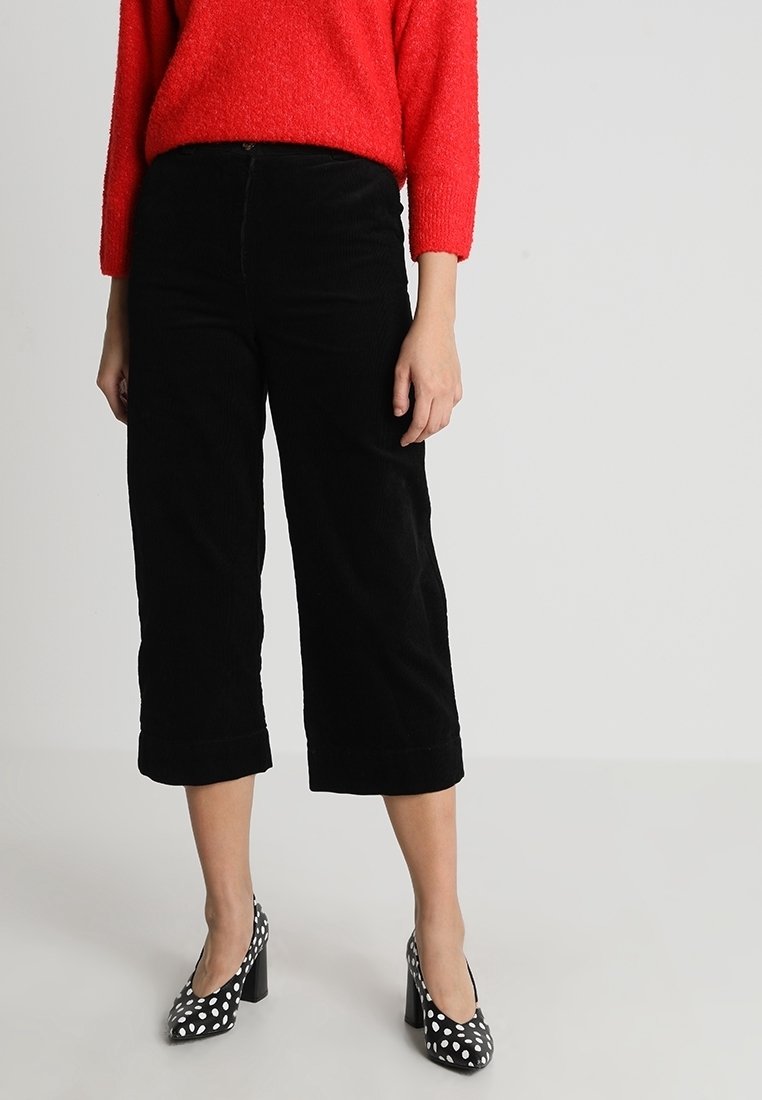 Culottes en velours côtelé noir avec un ourlet raccourci et des revers pliés, associées à un pull texturé rouge vif et des talons noirs à pois.
