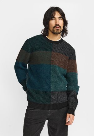 Mand med skulderlangt mørkt hår iført en multifarvet patchwork sweater og sorte jeans, stående mod en ensfarvet baggrund.