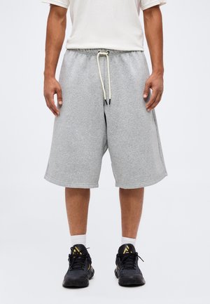 Homme portant un short gris clair mi-long avec cordon, chaussettes blanches, chaussures de sport noires et dorées, debout devant un fond uni.