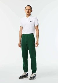 Camisa polo blanca con el logo de Lacoste, combinada con pantalones jogger verdes con cintura elástica y puños, llevada con zapatillas negras y blancas.