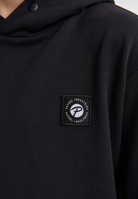 Felpa nera con cappuccio realizzata in tessuto morbido, dotata di una tasca frontale e di un logo quadrato con testo e design bianco sul lato sinistro.