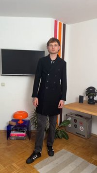 Čierny dvojradový kabát so štíhlym strihom, nosený cez čierno-bielo pruhovanú košeľu a šedé nohavice, doplnený čiernymi teniskami.