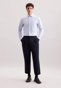 Lichtblauw overhemd met lange mouwen en knopen, voorzien van een borstzak; gecombineerd met donkerblauwe geplooide broek en zwarte schoenen. Glad stoftextuur.