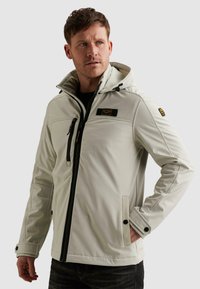 Mann trägt eine hellbeige Kapuzenjacke mit Reißverschluss in Schwarz und Brusttasche, schaut zur Seite, eine Hand in der Tasche.