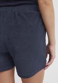 Mørkeblå frottéshorts med elastisk midje, som har en myk tekstur og en rett kant, vist fra baksiden med en hånd på siden.