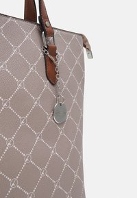 Sac fourre-tout en matériau texturé beige clair avec un motif en diamant, doté de poignées en cuir marron et d'une étiquette en métal argenté attachée par une chaîne.