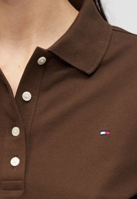 Καφέ μπλουζάκι πόλο με υφή, λευκά κουμπιά και μικρό κόκκινο, λευκό και μπλε ορθογώνιο λογότυπο στο στήθος.