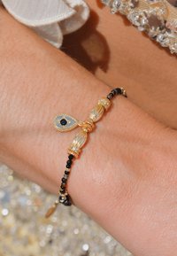 Bracelet en perles dorées et noires avec pendentif en forme de goutte, comprenant une pierre noire centrale et des accents décoratifs dorés. Texture lisse, design élégant.