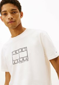 T-shirt bianco in cotone a maniche corte, con stampa in outline nera che riporta "TOMMY JEANS" e "NYC 1985."
