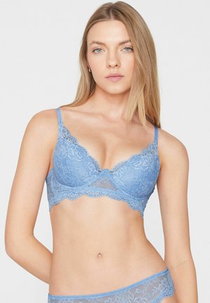 Junge Frau mit langen blonden Haaren, die einen hellblauen floralen Spitzen-BH und passende Spitzenunterwäsche vor einem einfarbigen Hintergrund trägt.