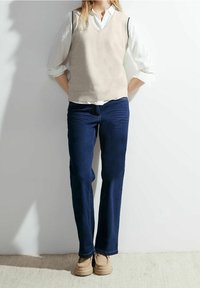 Beige gestrickte Weste über einem weißen Hemd mit Kragen, kombiniert mit dunkelblauen Jeans und hellbraunen Schuhen. Der Look hat eine lässige Ästhetik.