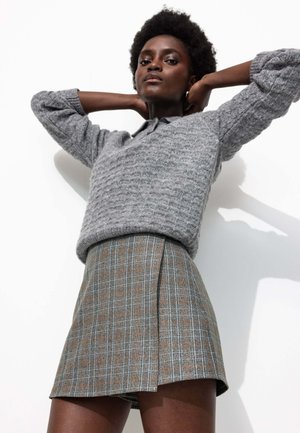 Pull en tricot gris avec un motif en relief, associé à une jupe mini à carreaux dans des tons gris et marron, présentant un design plissé.