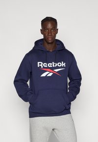 Hanorac bleu marin cu buzunar frontal, având un logo Reebok alb și roșu. Material moale, glugă ajustabilă cu șnur și manșete texturate.