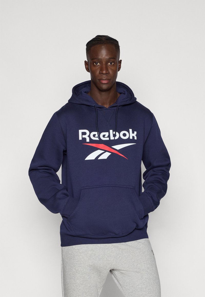 Hanorac bleu marin cu buzunar frontal, având un logo Reebok alb și roșu. Material moale, glugă ajustabilă cu șnur și manșete texturate.