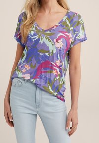 Lila V-kaula T-paita trooppisella kukkakuviolla, jossa on vaaleanpunaista, sinistä ja vihreää, valmistettu kevyestä kankaasta; yhdistettynä vaaleisiin farkkuihin.