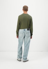 Mann som vender bort, iført en olivenfarget langermet skjorte, lyseblå løse jeans og hvite og grå sneakers mot en enkel hvit bakgrunn.