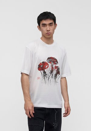 Ung mand iført en hvid T-shirt med en rød og sort vandmandsgrafik, stående foran en ensartet lys grå baggrund.
