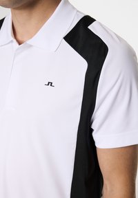 Vit polo skjorta med svarta sidopaneler, korta ärmar och krage. Inkluderar en liten svart logotyp på vänstra bröstet. Slät textur.