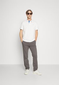 Weißes Polohemd mit blauen Kragenakzenten, graue Hosen und weiße Sneaker. Modell mit Sonnenbrille, steht vor einem neutralen Hintergrund.