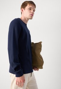 Lange mouw navy waffle-gebreide shirt, gecombineerd met crèmekleurige broek, terwijl hij een grote beige tas met een textuur vasthoudt.