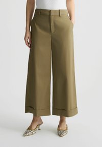 Pantaloni a gamba larga verde oliva con zip frontale, tasche laterali e orli cuffiati, realizzati in tessuto elastico, abbinati a tacchi con motivo.
