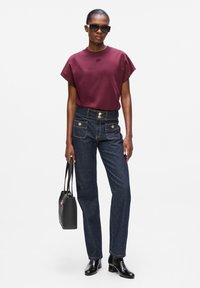 Burgunderfarbene Kurzarmbluse mit geprägtem Detail, kombiniert mit hochgeschnittenen, dunklen Denim-Jeans mit goldenen Knopfakzenten und zwei vorderen Taschen.