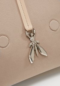 Bežová plátěná taška s kovovým šarmem ve tvaru hmyzu připevněným na koženém popruhu. Textura je hladká s vyvýšenými tečkovanými detaily na povrchu.