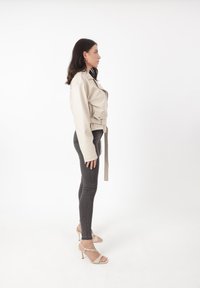 Giacca beige cropped con vita belted, tasche laterali e colletto strutturato, abbinata a jeans neri aderenti e tacchi con cinturini.