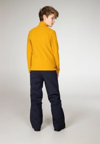 Pullover in fleece giallo con colletto alto, abbinato a pantaloni impermeabili blu scuro. L'outfit presenta una vestibilità comoda e una texture liscia.