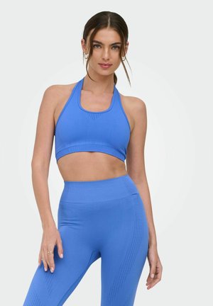 Vrouw met bruin haar draagt een blauwe naadloze halter sportbeha en high-waist legging, staand met één hand ontspannen langs het lichaam.