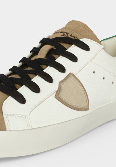 Gros plan sur une basket avec des empiècements beiges et blancs, des lacets noirs et un patch beige en forme de bouclier sur le côté.